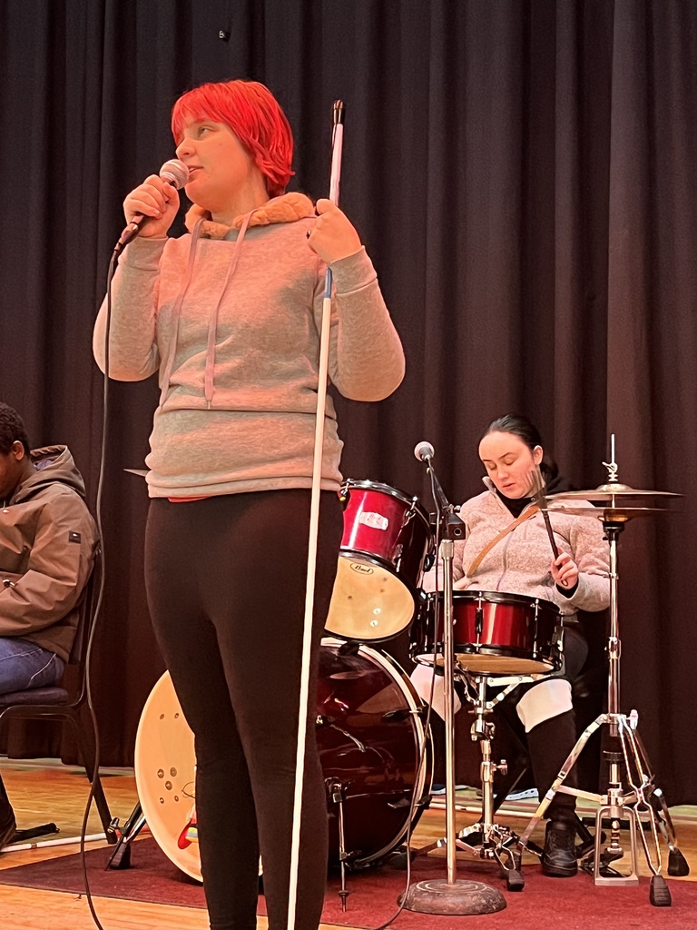 One student holding a microphone and her cane while singing, one student playing drums in the background 