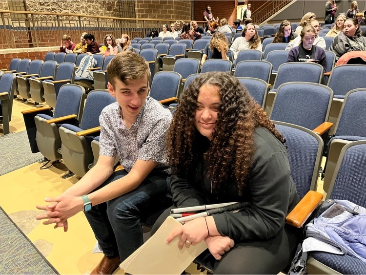 two students sitting in an auditorium with a few other students scattered in other seats 