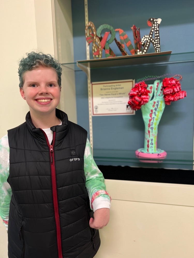 female student with short curly blue colored hair smiles standing next to her art piece. red colored foam pieces separated by a paper mache hand signify the two halves of the brain