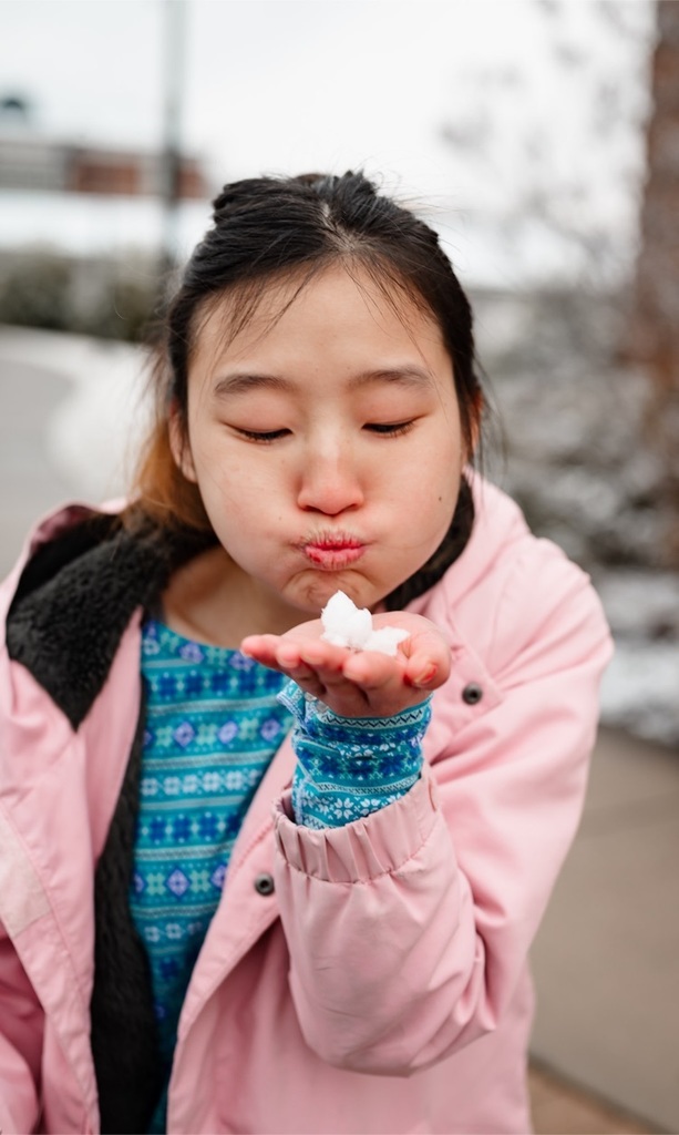 a girl holds a small clump of snow in one hand and holds it up to her face. her cheeks are puffed out, ready to blow the snow