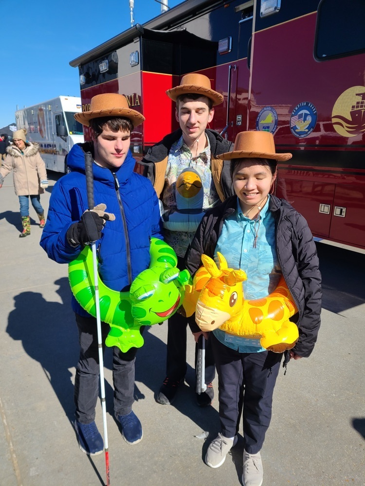 three young adult students wearing inflatable inner tubes designed as a frog, a giraffe, and a bird. each student is also wearing a wide brimmed hat. two students are holding canes  