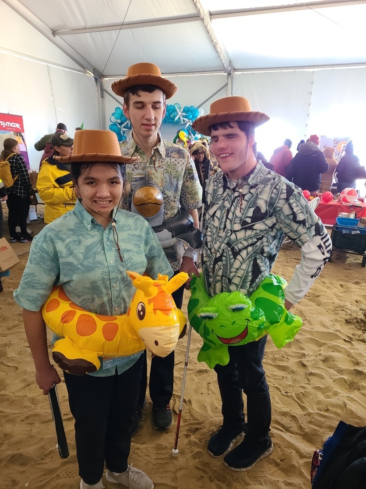 three young adult students wearing inflatable inner tubes designed as a frog, a giraffe, and a bird. each student is also wearing a wide brimmed hat. two students are holding canes