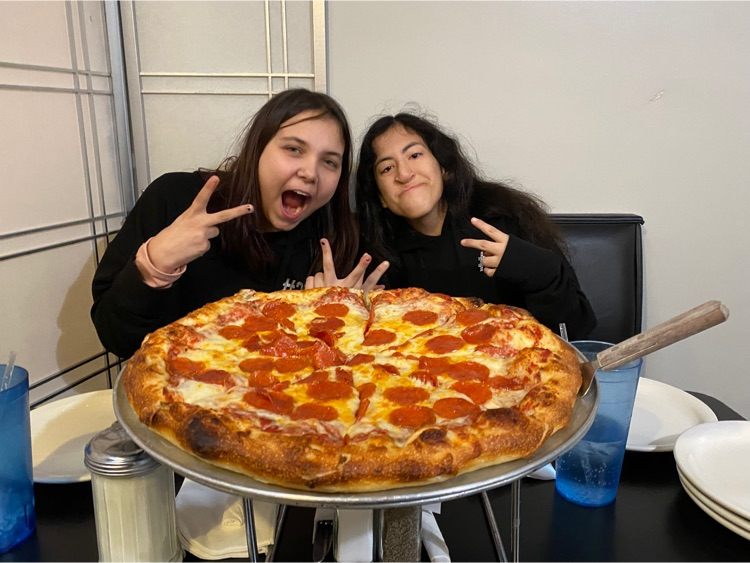 two girls sit behind a large pizza and hold up peace signs