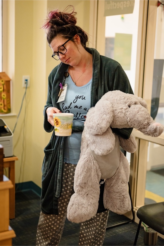 female staff wearing pajamas pretends to sleep standing up while wearing pajamas and holding a stuffed elephant and a coffee cup