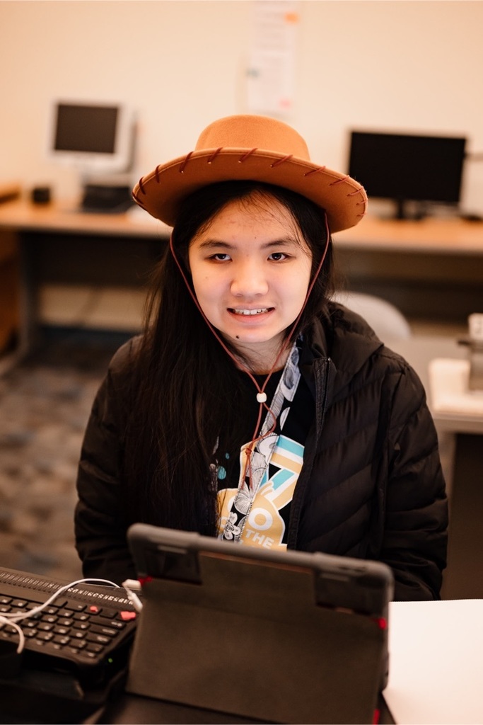 female student wearing a wide brimmed hat smiling while sitting at a desk 