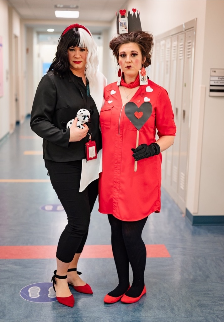 two female staff standing in a hallway. one dressed as cruella de’vil and one dressed as the queen of hearts. both with mean expressions  