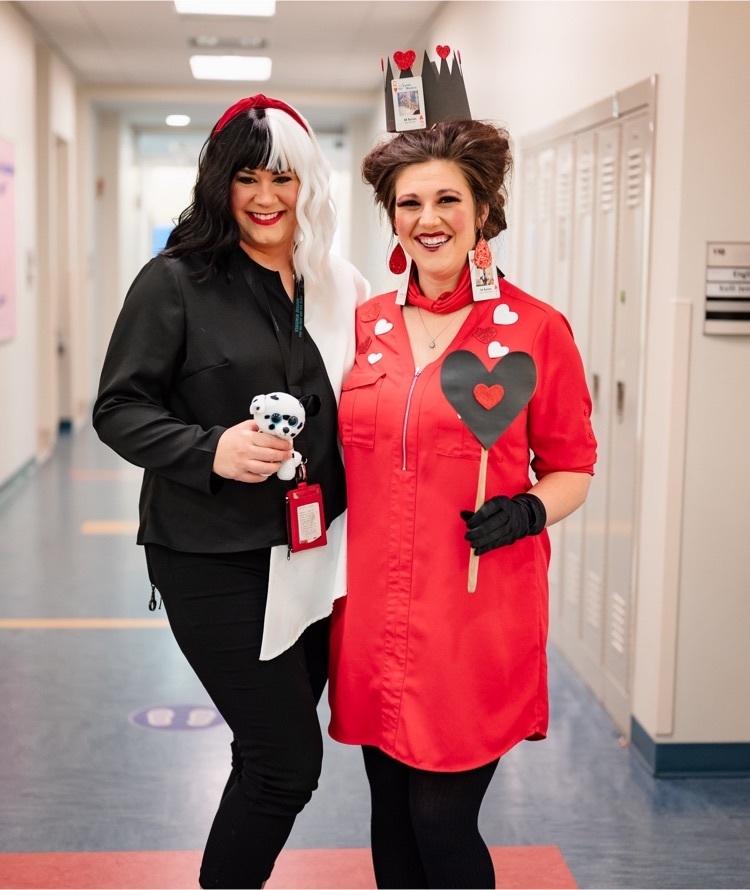 two female staff standing in a hallway. one dressed as cruella de’vil and one dressed as the queen of hearts. both smiling  