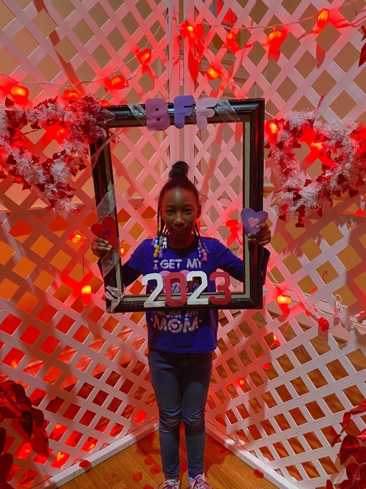 elementary student holding a frame that says BFF 2023 and standing in front of a backdrop decorated with hearts and red lights