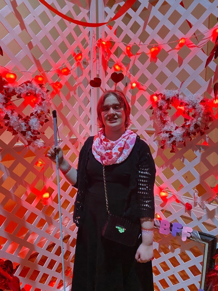 high school student wearing a black dress with a valentines scarf and a heart headband standing in front of a backdrop decorated with hearts and red lights 