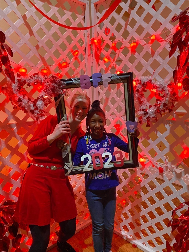elementary student and female staff holding a frame that says BFF 2023 standing in front of a backdrop decorated with hearts and red lights 