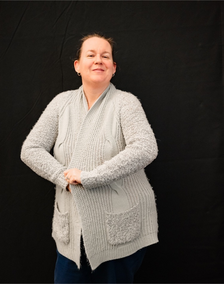 female staff stands against a black background wrapped in a fuzzy gray sweater 