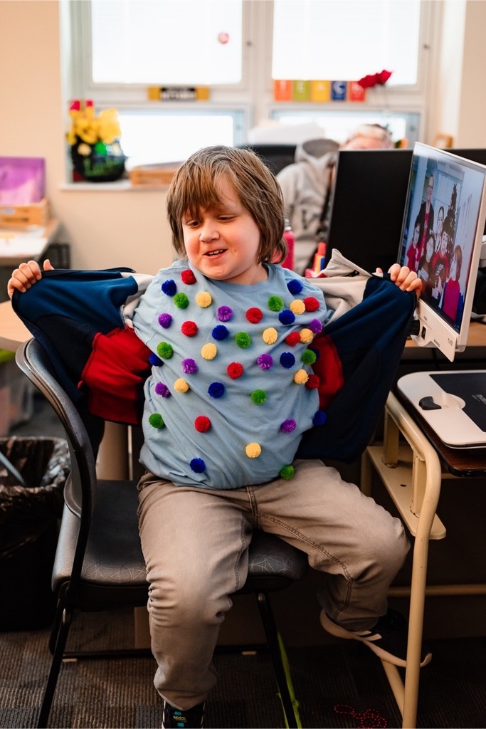 male student sits in a classroom showing his T-shirt adorned with many colorful fuzzy pompoms 