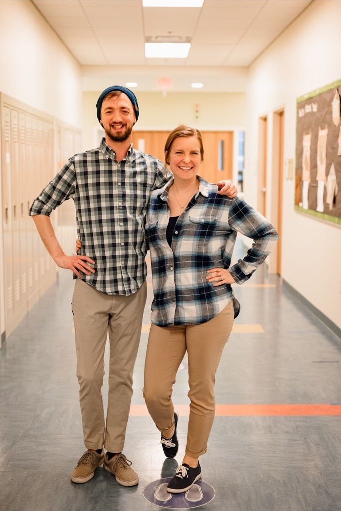 one female and one male staff wearing blue plaid shirts stand together in the hallway smiling at the camera