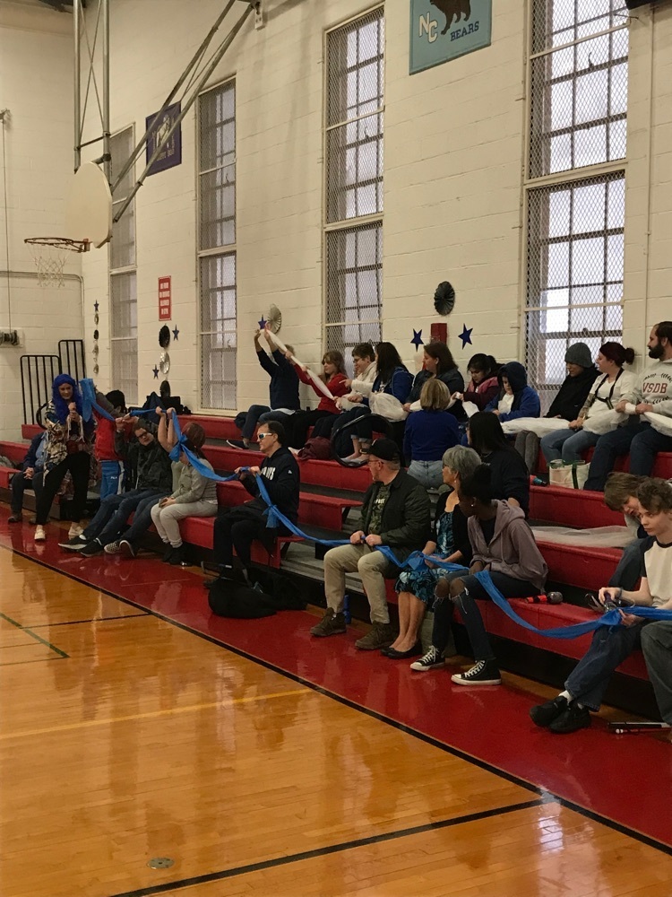 group of students and staff sitting on the bleachers holding onto a long piece or fabric