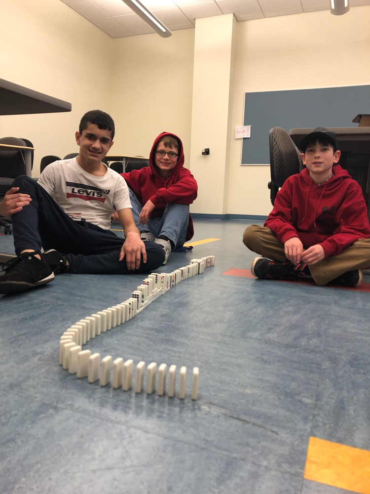 three male students sitting on the floor by a line of dominos