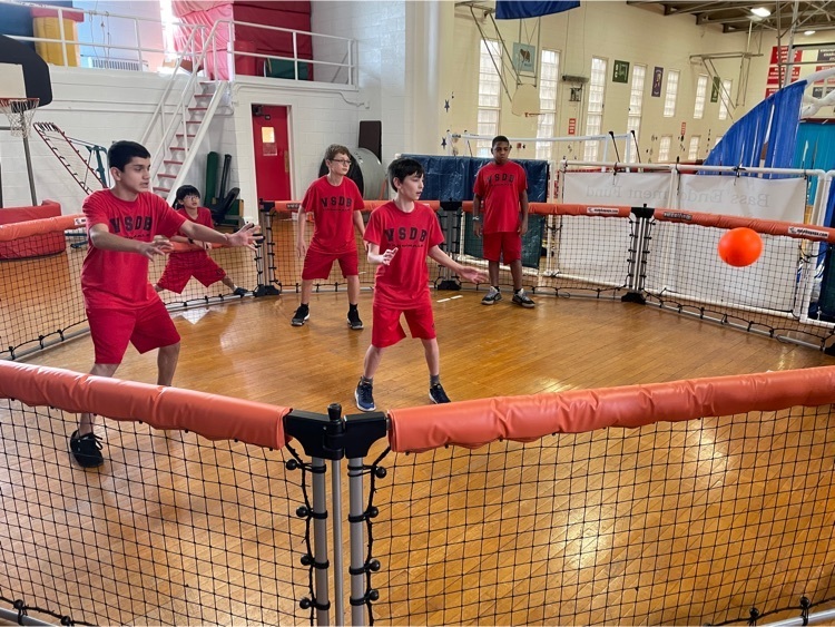 7 young students stand inside a fenced circle playing with a red ball
