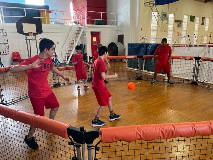7 young students stand inside a fenced circle playing with a red ball