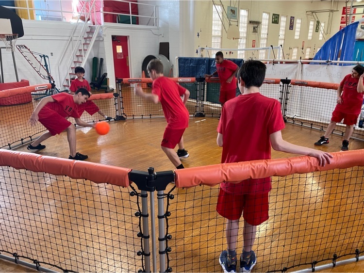 7 young students stand inside a fenced circle playing with a red ball