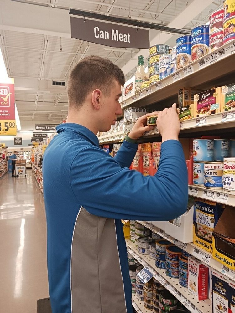 male student wearing a food lion uniform arranges a box on a shelf in the grocery store