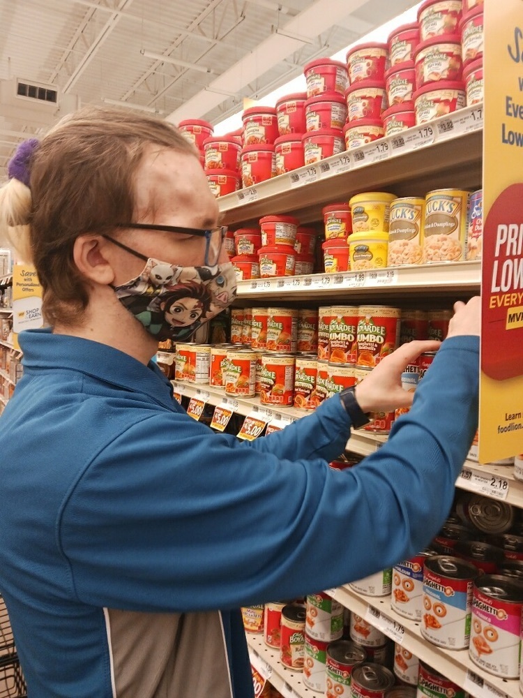 male student wearing a food lion uniform arranges a can on a shelf in the grocery store
