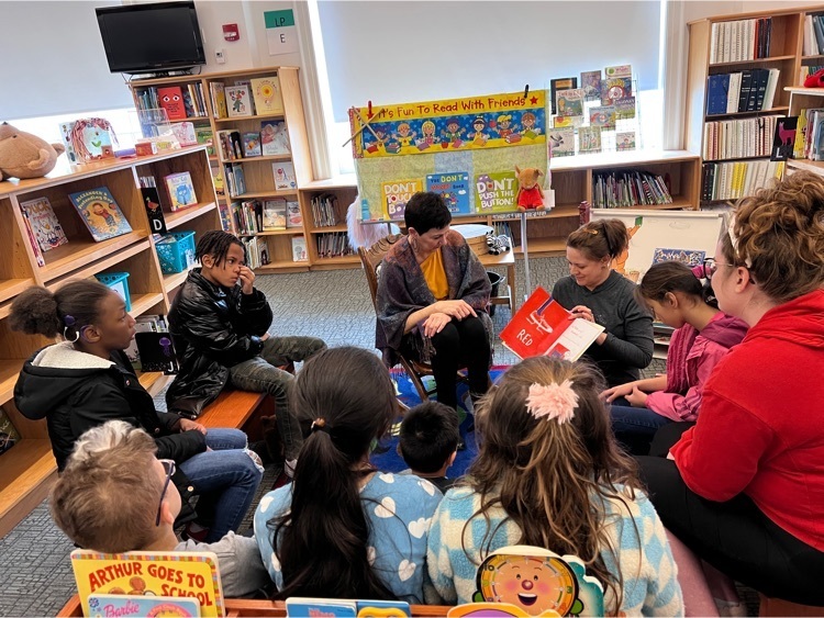 two female staff sit among a group of elementary students reading a story