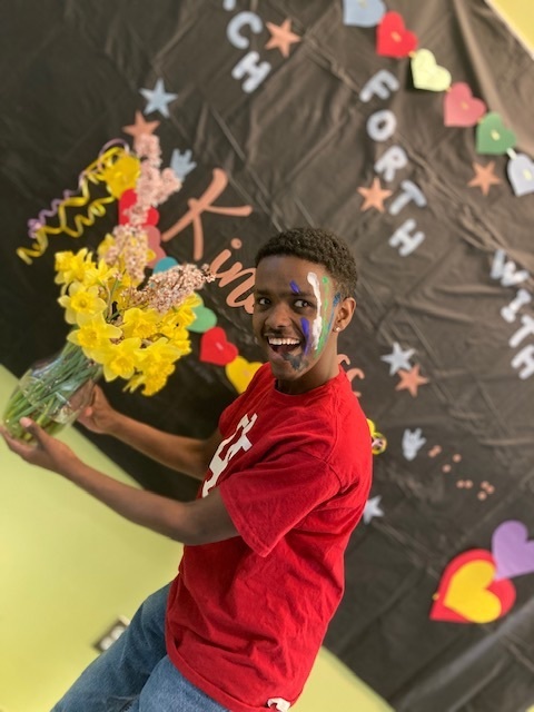 male student posing with a vase of yellow flowers. the kindness week photo backdrop is behind him