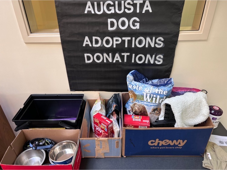 sign with white letters on a black background says Augusta dog adoptions donations. three boxes with food, bowls, and toys for dogs sit on a table 