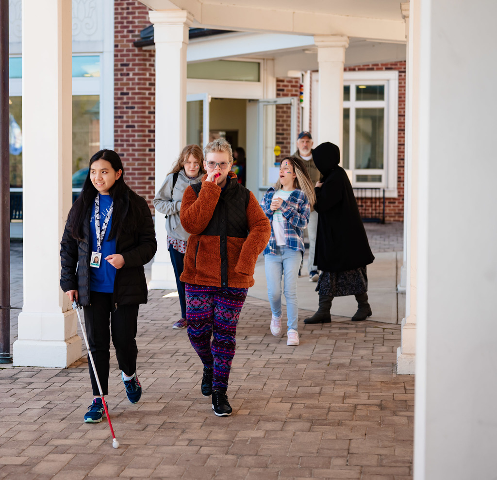 four female students walk along a walkway. one has a white cane, one is blowing a whistle, and two are laughing 