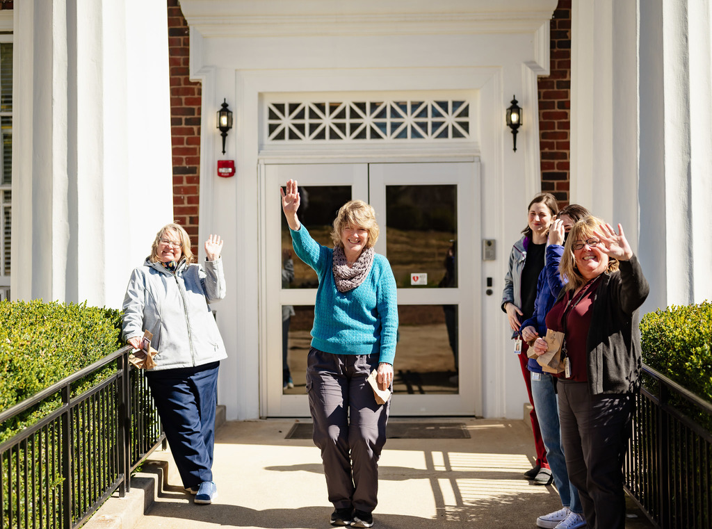 four female staff from human resources stand outside their building waving at the parade of students going by. the director of human resources holds a bag of candy that was given to their office.