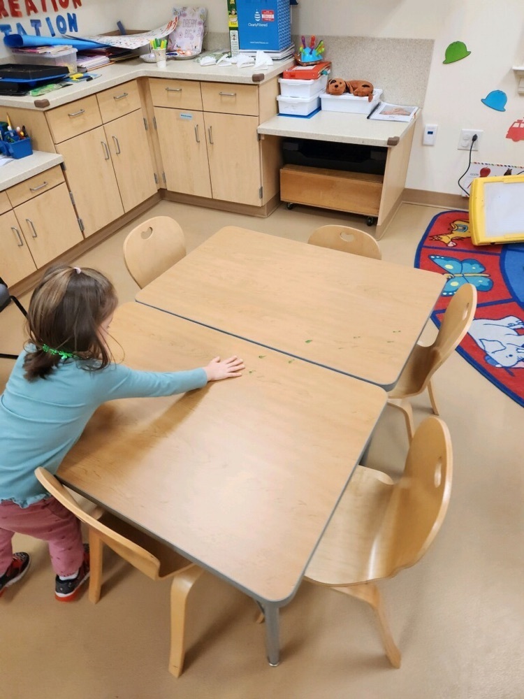 green “leprechaun footprints” on the tables in the pre-K classroom and a female pre-k student leaning over the table to point at and look at them 