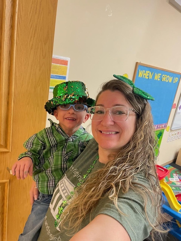 one male pre-k student being held by one female staff. they are both wearing green shirts and the boy has a sequined green hat on