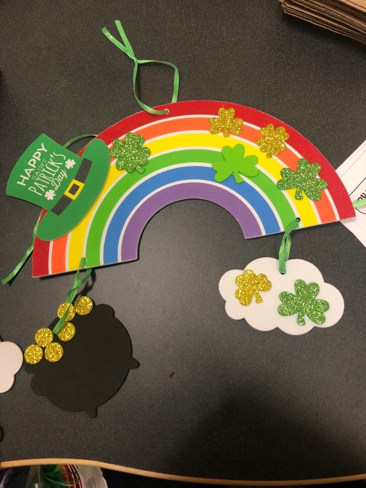 a foam craft of a rainbow with glittery green shamrocks stuck to it and a green hat that says happy st Patrick’s day. at the end of one side of the rainbow is a pot of gold. the other side has a white cloud with green shamrocks attached to it 