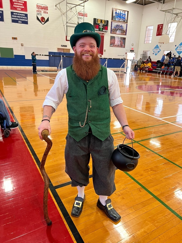 male staff dressed in a full leprechaun outfit. he has a natural red beard, a black hat, a green vest over a long sleeve white shirt, black trousers, black shoes with gold buckles, a cane, and a black pot full of “gold"