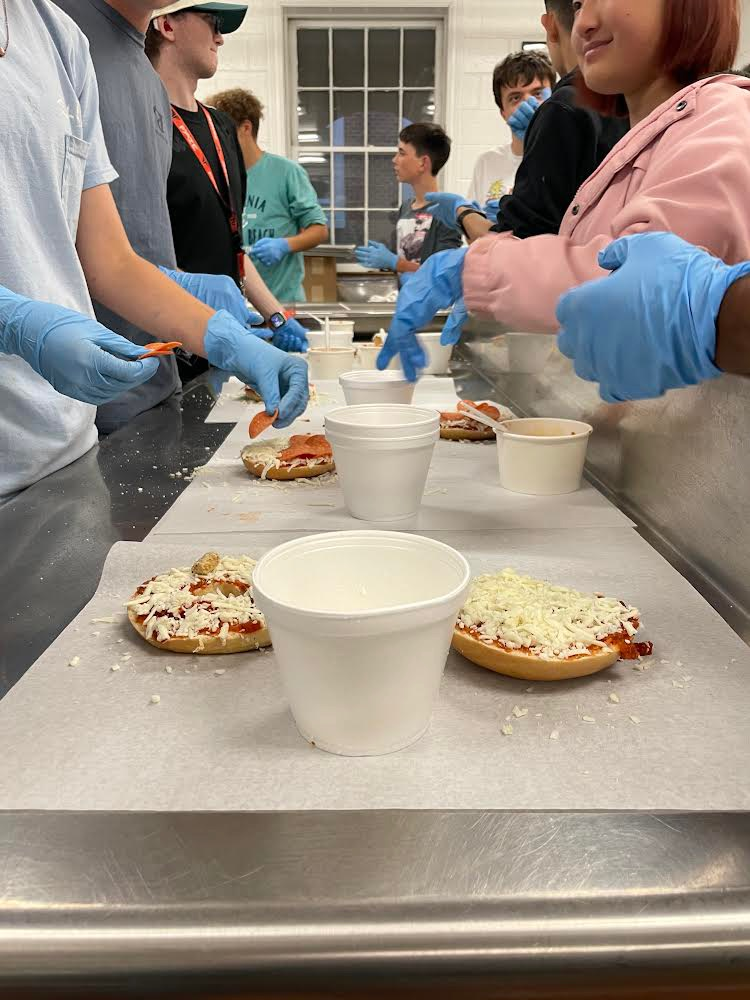 closeup of pizza bagels and students’ gloved hands putting on the toppings